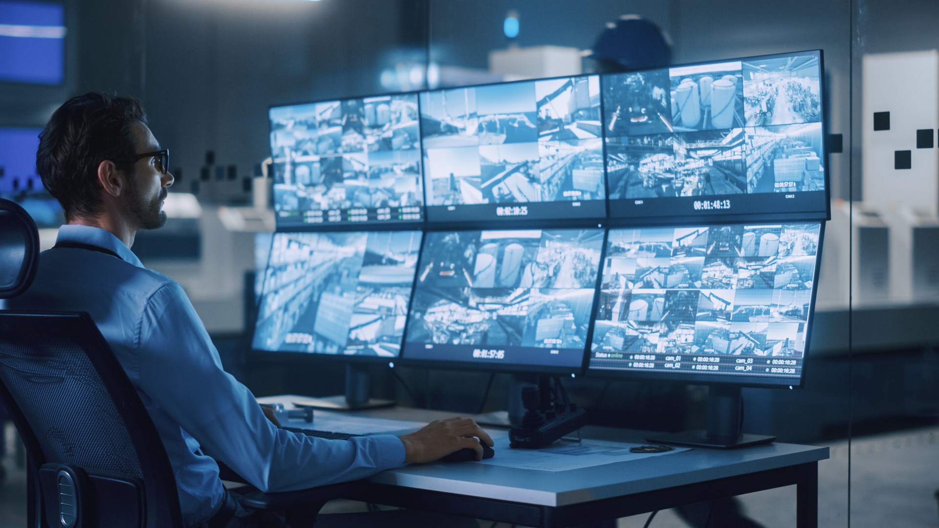 Industry 4.0 Modern Factory: Security Operator Controls Proper Functioning of Workshop Production Line, Uses Computer with Screens Showing Surveillance Camera Feed. High-Tech Security